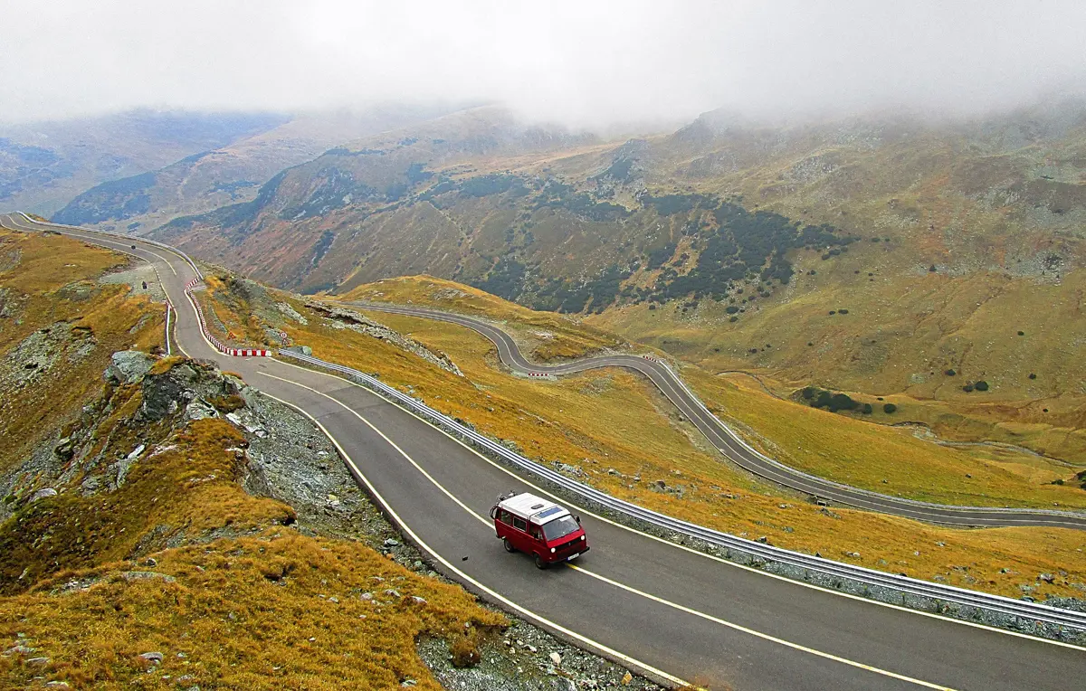 A campervan driving at the top of a long, windy mountain road with a scenic view behind
