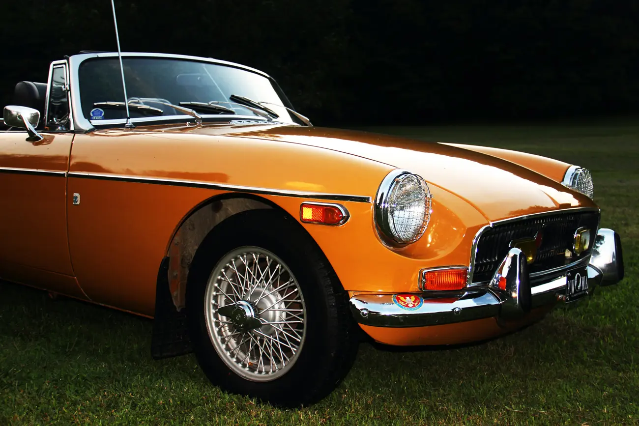 An orange MGB roadster parked on grass with its roof down at dusk