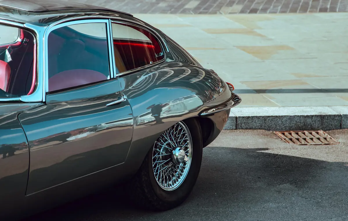 Th rear corner of a dark grey Jaguar E-Type with red interior parked in-front of a curb