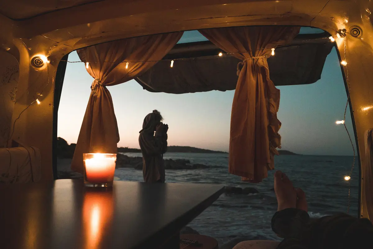 A candle on a table in a campervan as someone lays down looking at the open rear door at dusk