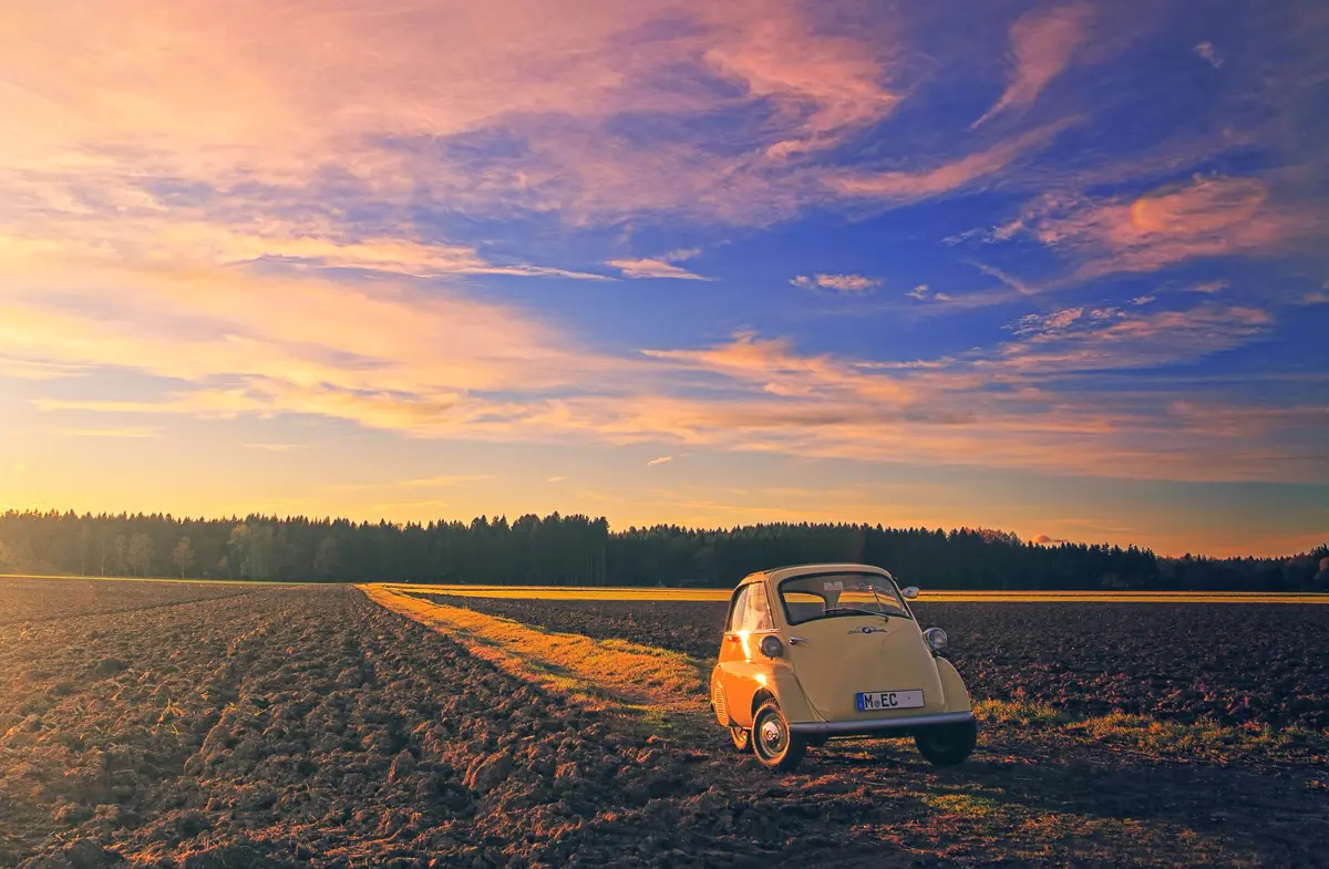 An Isetta bubble car parked on a dirt track through a large field at sunset